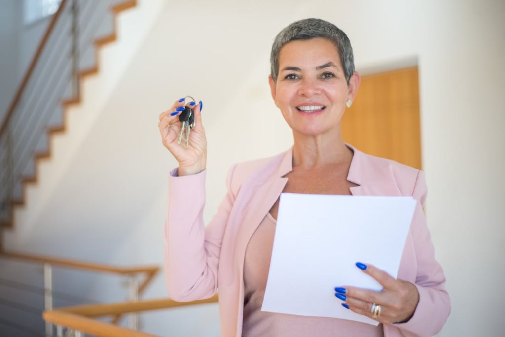 Confident woman in a pink blazer holding keys and a document, smiling indoors.